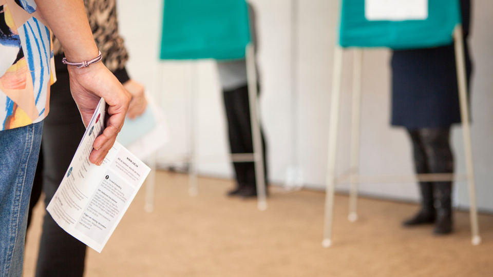 Foto som visar en vallokal där några står i kö för att rösta och några står bakom en skärm och röstar. 